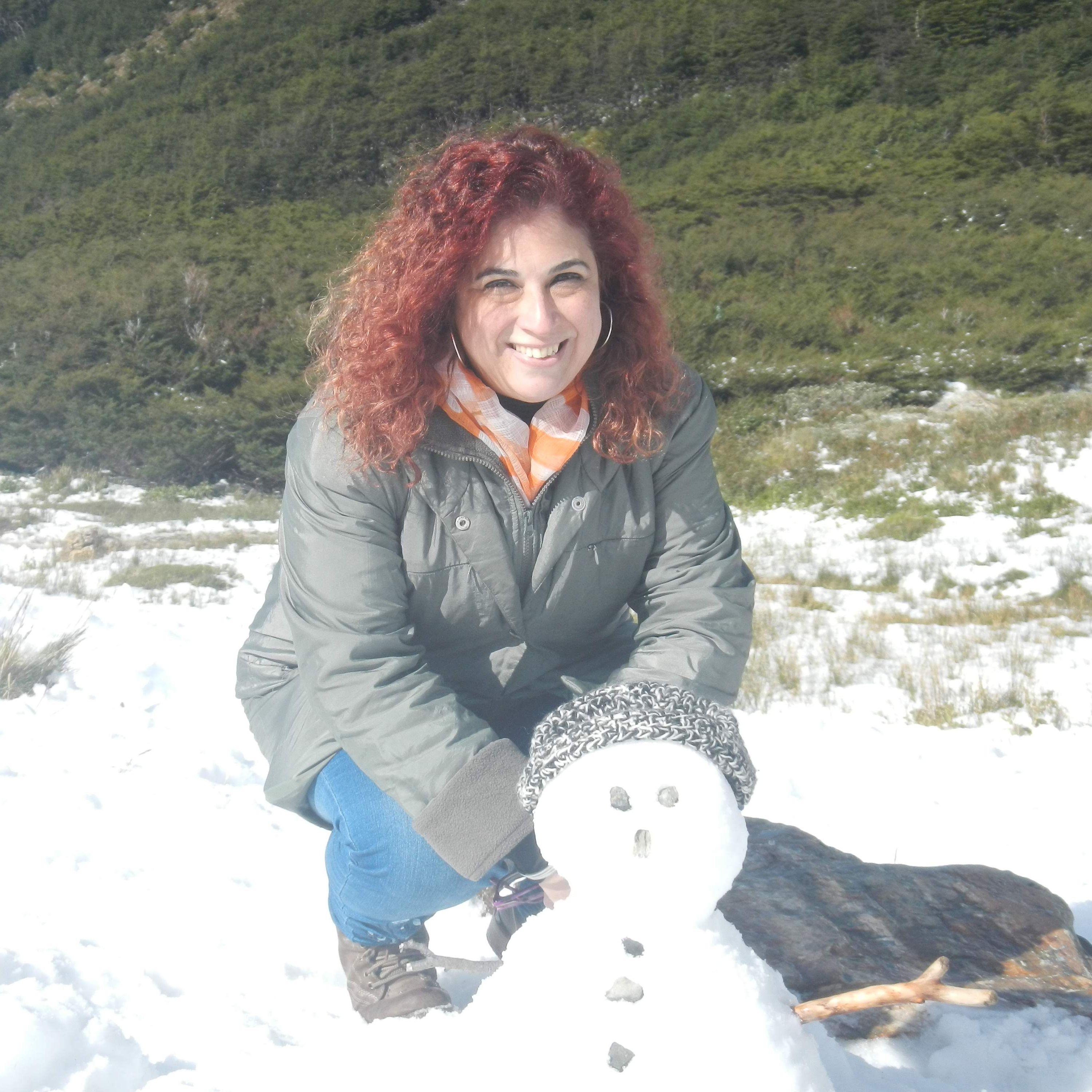 Ana Saladino, tallerista de Ciempiés. Ana tiene cabellos cobrizos, rizados y largos hasta los hombros. En la foto está agachada posando junto a un pequeño muñeco de nieve.