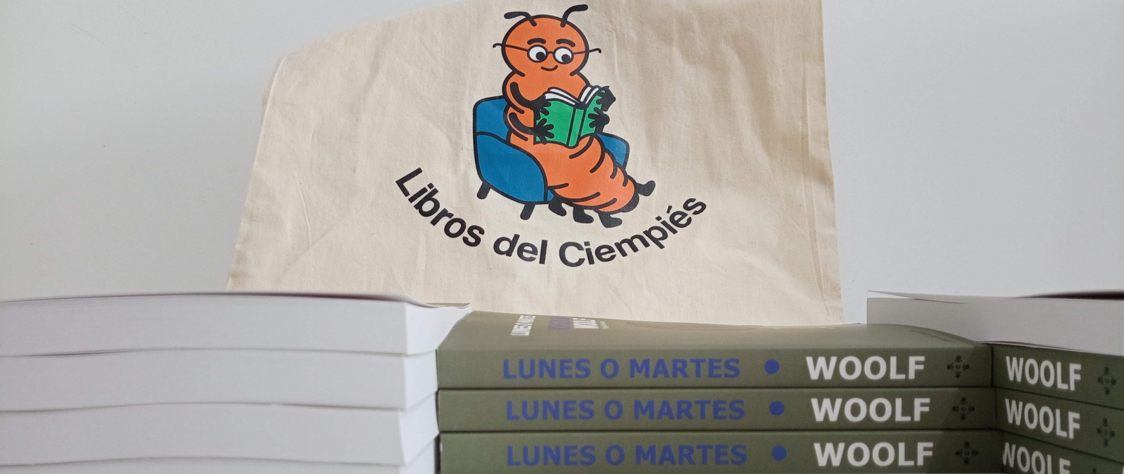 Una tete bag color crudo con la imagen del »Ciempiés lector» debajo de la ilustración dice «Libros del Ciempiés». La bolsa está por detrás de una pila de ejemplares del libro «Lunes o martes», de Viriginia Woolf, editado por Libros del Cimpiés para su colección «Clásicos Accesibles».