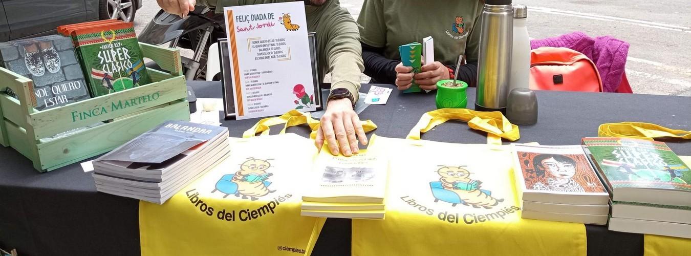 En la foto se ve la parada que Ciempiés en Sant Jordi 2024. Sobre una mesa cubierta con un mantel negro se observan varios libros y un par de tote bags amarillas de "Libros del Ciempiés".  